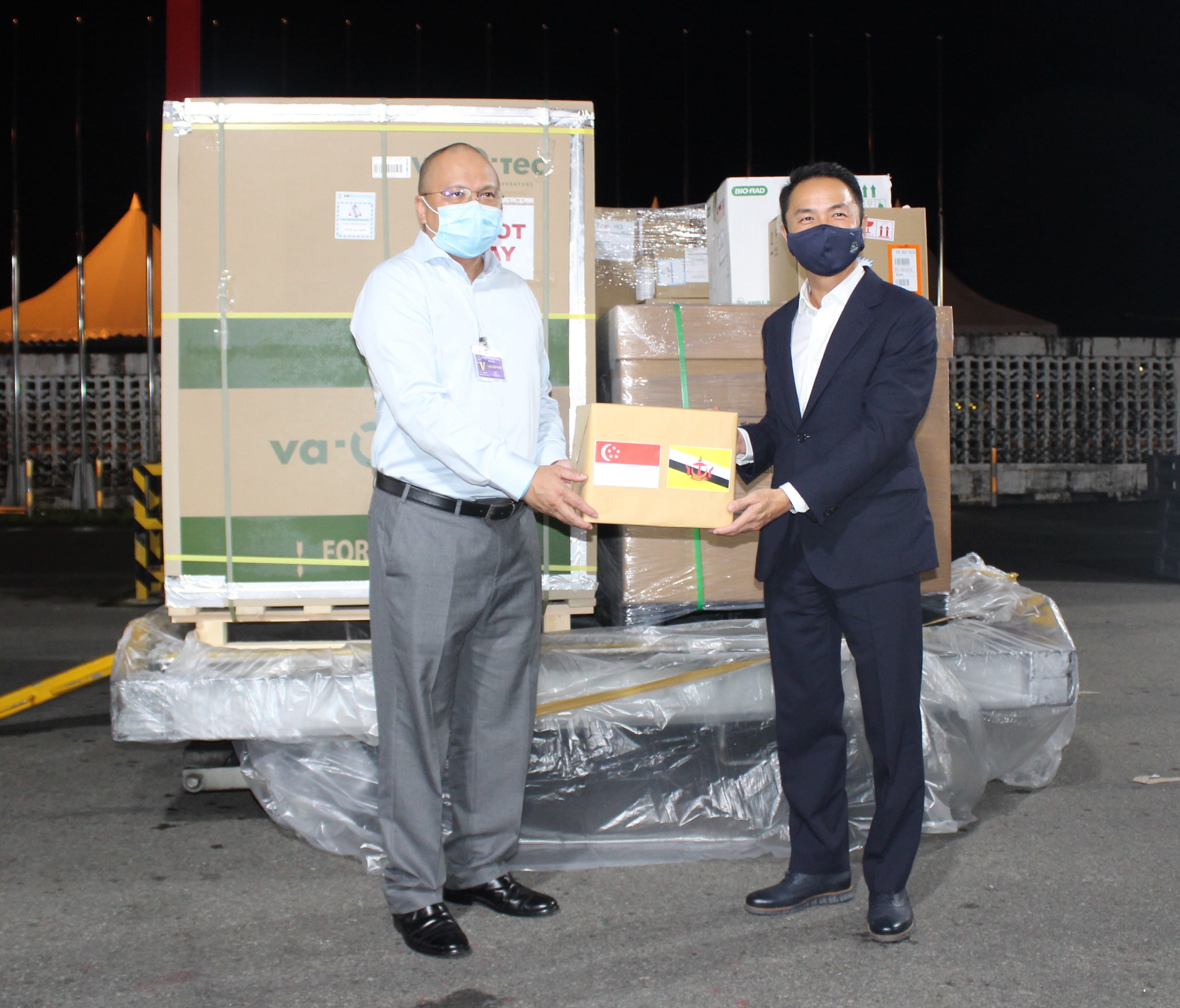 Two men wearing face masks hold a box with Singapore and Brunei flags in front of cargo.